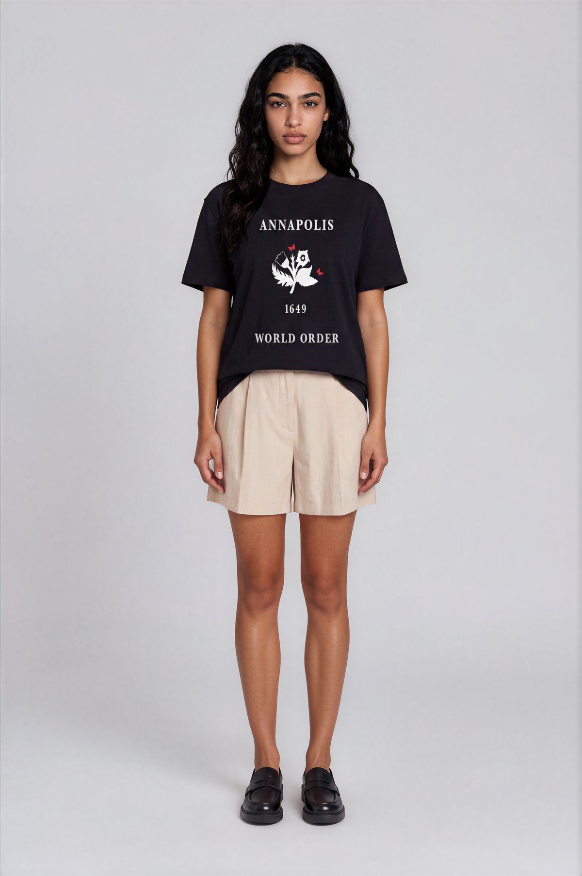 A model stands against a light gray studio background wearing a black graphic T-shirt with white text reading 'ANNAPOLIS 1649 WORLD ORDER' and a central emblem, paired with light beige shorts and black loafers.