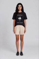 A model stands against a light gray studio background wearing a black graphic T-shirt with white text reading 'ANNAPOLIS 1649 WORLD ORDER' and a central emblem, paired with light beige shorts and black loafers.