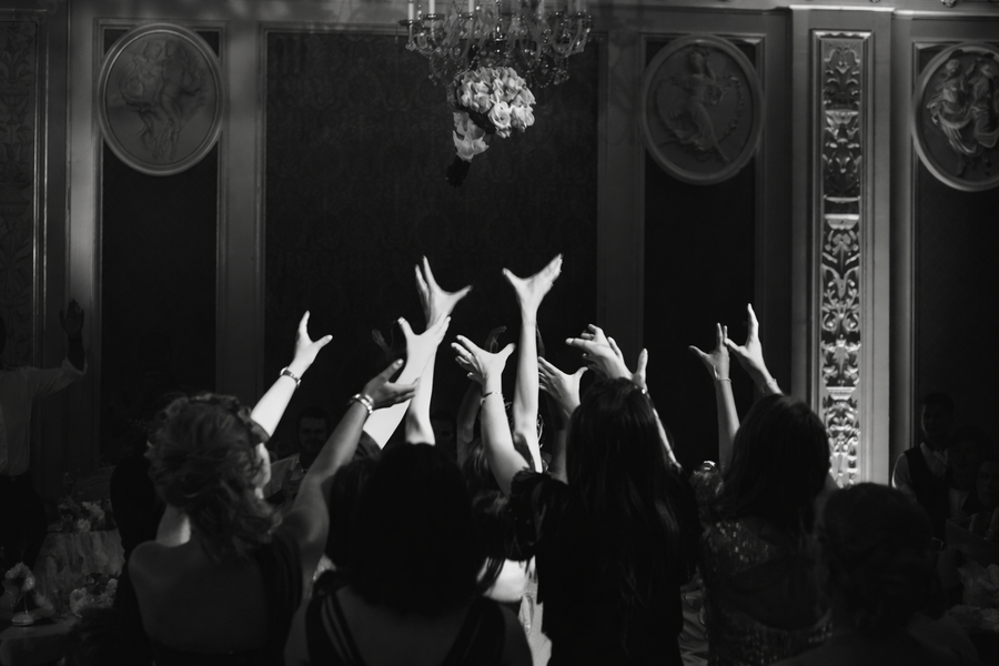 Anonymous hands reaching for a thrown bouquet under a chandelier — black and white ballroom photograph