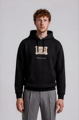 A man wearing a black pullover hoodie with a beige graphic print on the chest and gray trousers, standing against a light gray studio background.