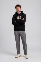 A man modeling a black pullover hoodie with a beige graphic on the chest, paired with gray trousers and white sneakers, standing against a light gray studio background.