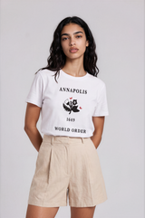 A woman modeling a white graphic t-shirt with a black owl design and text, paired with beige pleated shorts, against a light gray studio background.